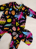 Stranger Things Romper and Hat Set by Little Loves Cornwall, bold abstract multicolour print on black, shown on hanger with tropical leaf background