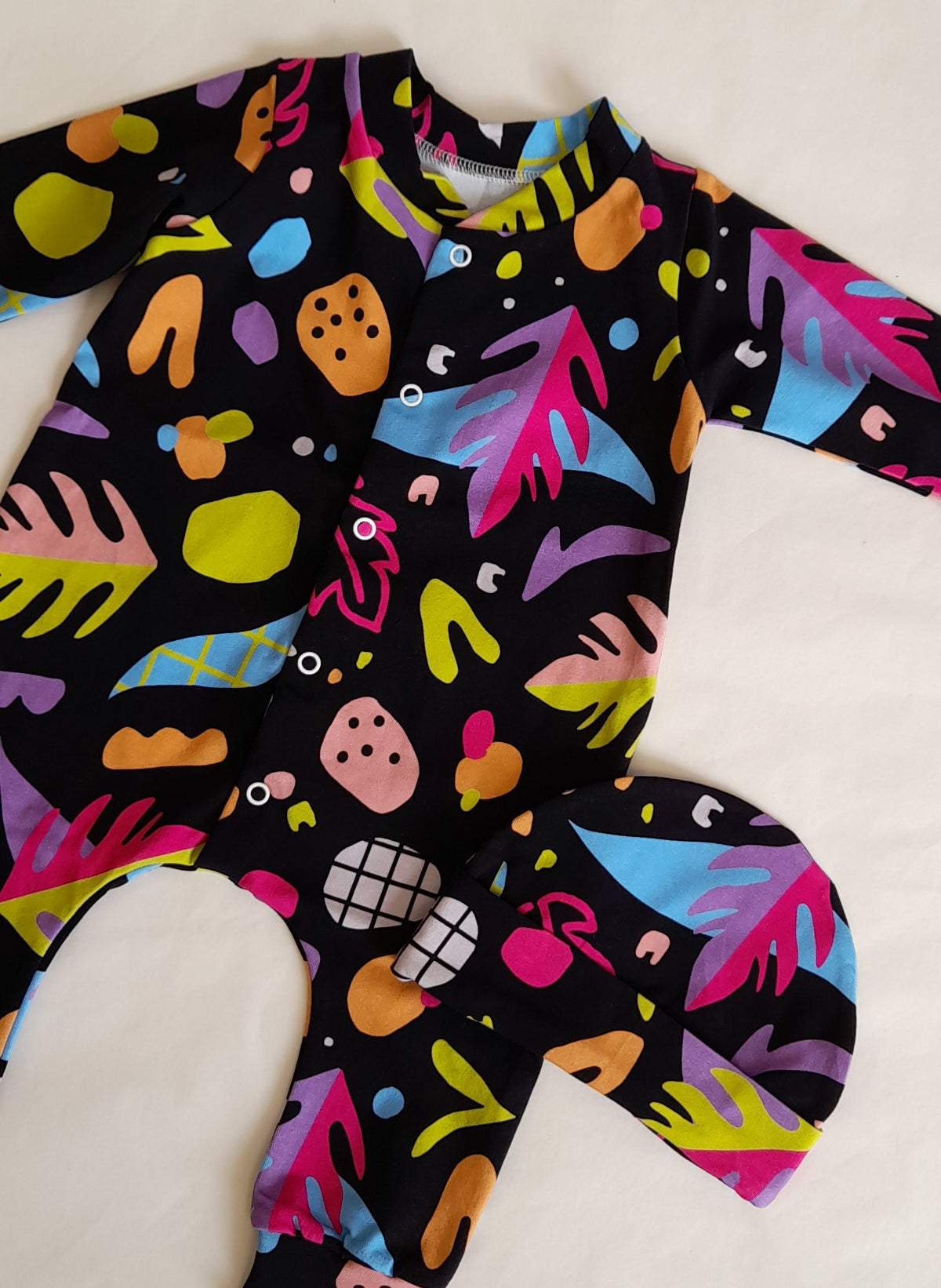 Stranger Things Romper and Hat Set by Little Loves Cornwall, bold abstract multicolour print on black, shown on hanger with tropical leaf background